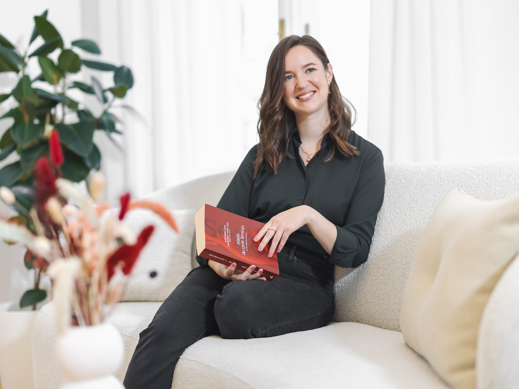 Eine Frau sitzt entspannt auf deinem Sofa. Sie lächelt und strahlt eine freundliche professionalität aus. Sie hält ein rotes Buch in der Hand. Es ist ein Buch über den weiblichen Zyklus